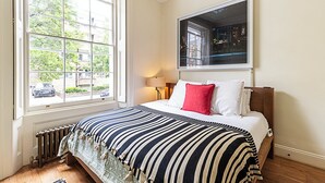 A bedroom with a large window, a bed with striped bedding, a red pillow, and a nightstand with a lamp. - Classic Luxury in Central London (London)