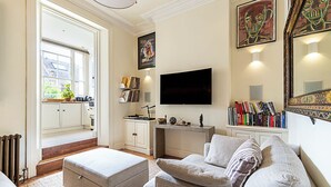 A living room with a sofa, ottoman, and a flat-screen TV mounted on the wall. There is a bookshelf with books and a small table with decorative items. - Classic Luxury in Central London (London)
