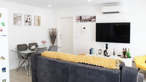 A modern living room with a sofa, a dining table, chairs, a flat-screen TV, and a wall-mounted air conditioner. - Blossom Cozy Home (Ponta Delgada)