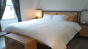 A neatly made bed with white linens, a wooden bench at the foot of the bed, a bedside table with a lamp, and a wall-mounted shelf above the bed. - Eclectic Decor 5 Br house, 2 Baths & 2 Kitchens with WiFi and AC in Old Forge  (Scranton)
