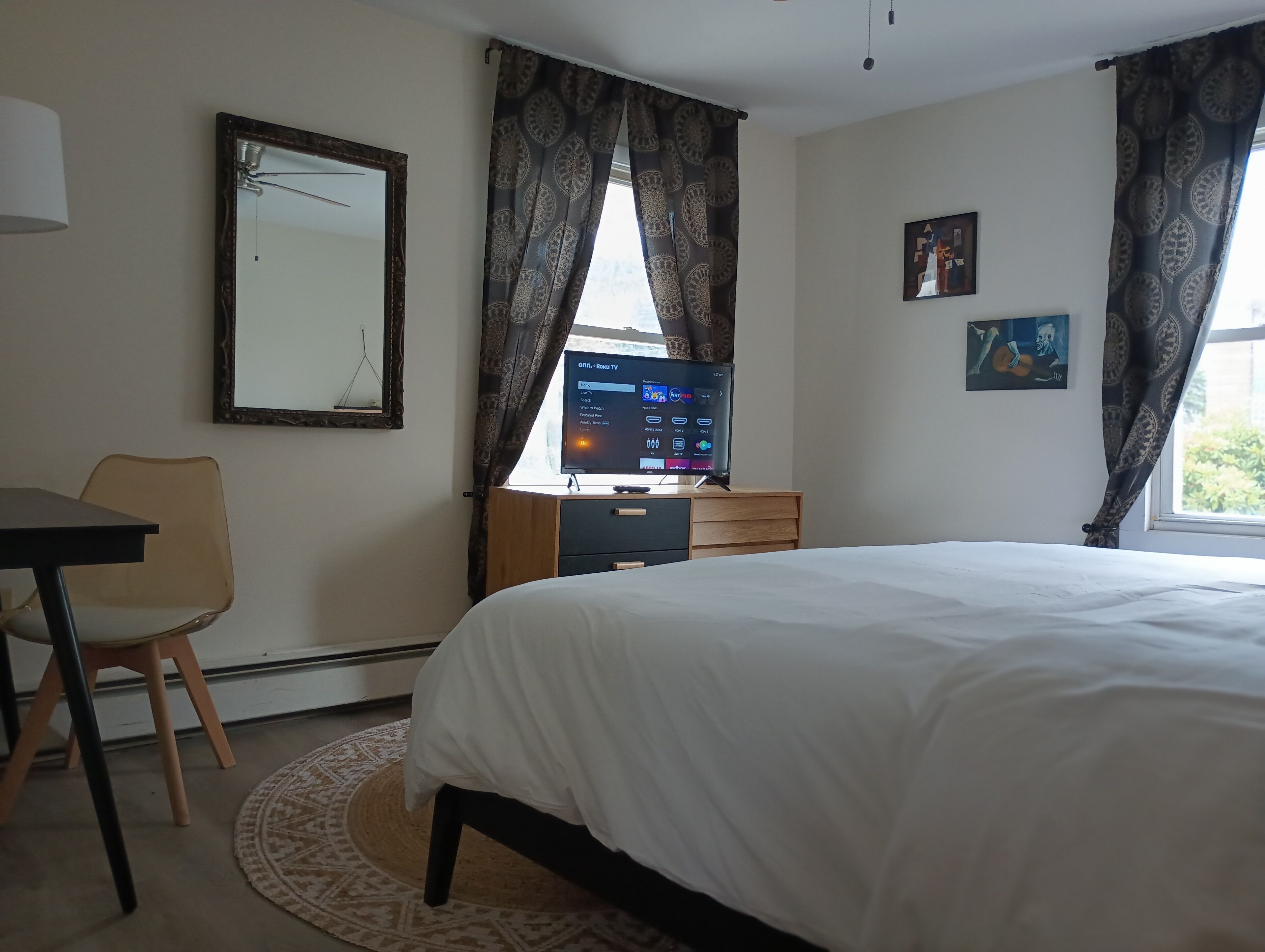 A bedroom with a bed, a desk with a computer, a chair, a window with curtains, and a painting on the wall.