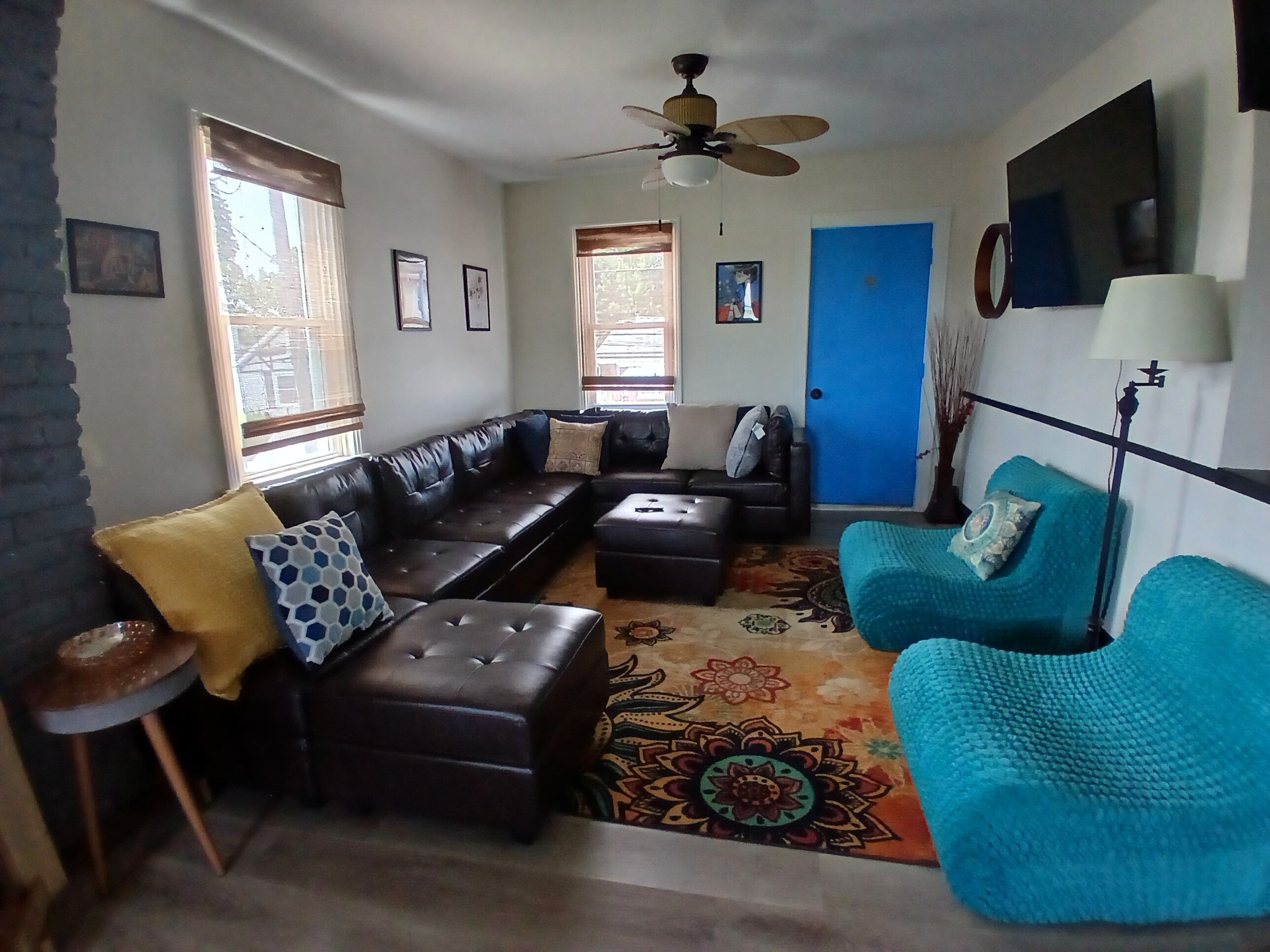 A living room with a sofa, ottoman, and a blue armchair. There is a ceiling fan, a TV mounted on the wall, and a door with a blue frame.
