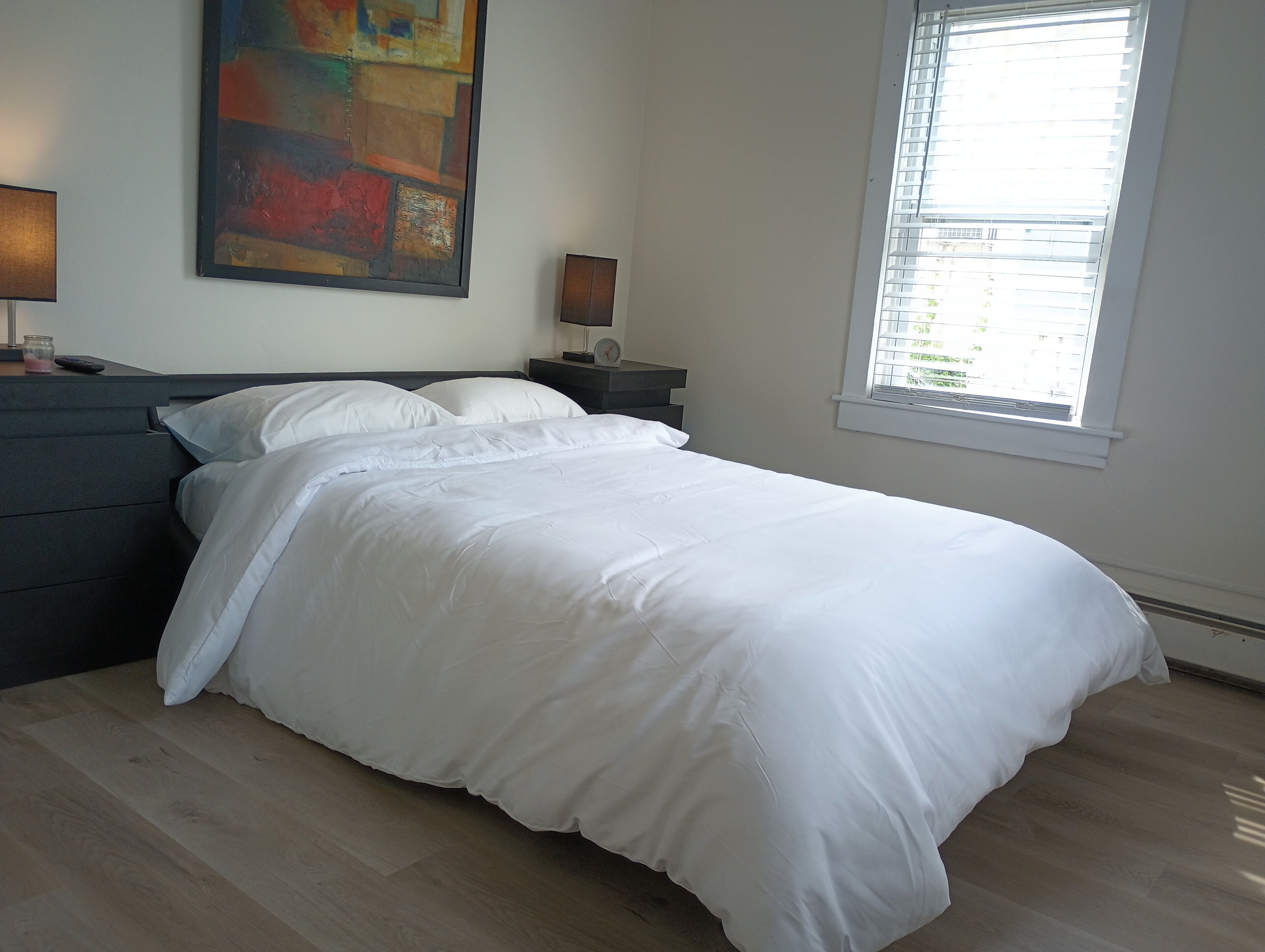 A neatly made bed with white linens in a room with a wooden floor, a painting on the wall, and a window with blinds.