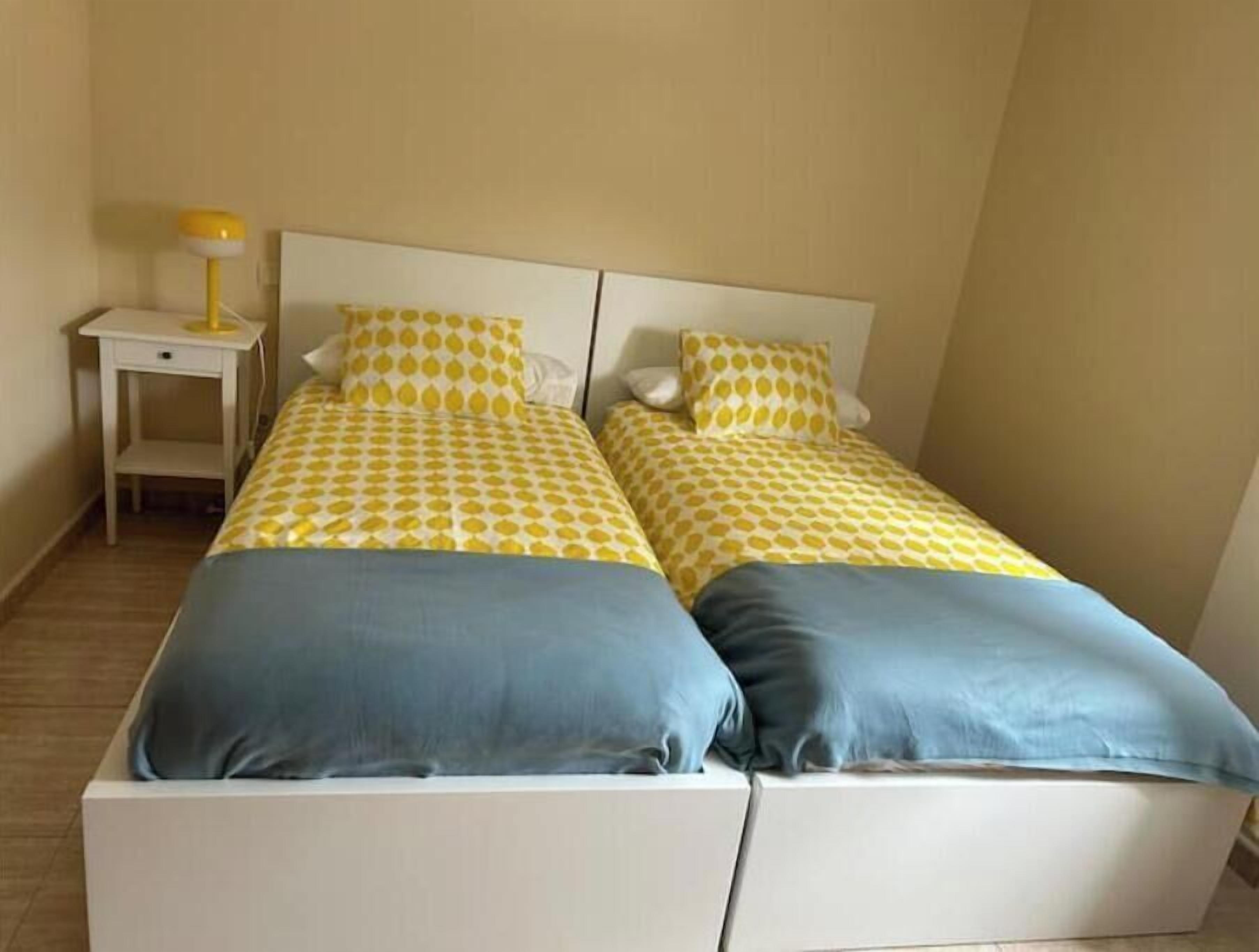 A double bed with a yellow and white patterned bedspread and two pillows, one blue and one grey, in a room with a white nightstand and a yellow lamp.