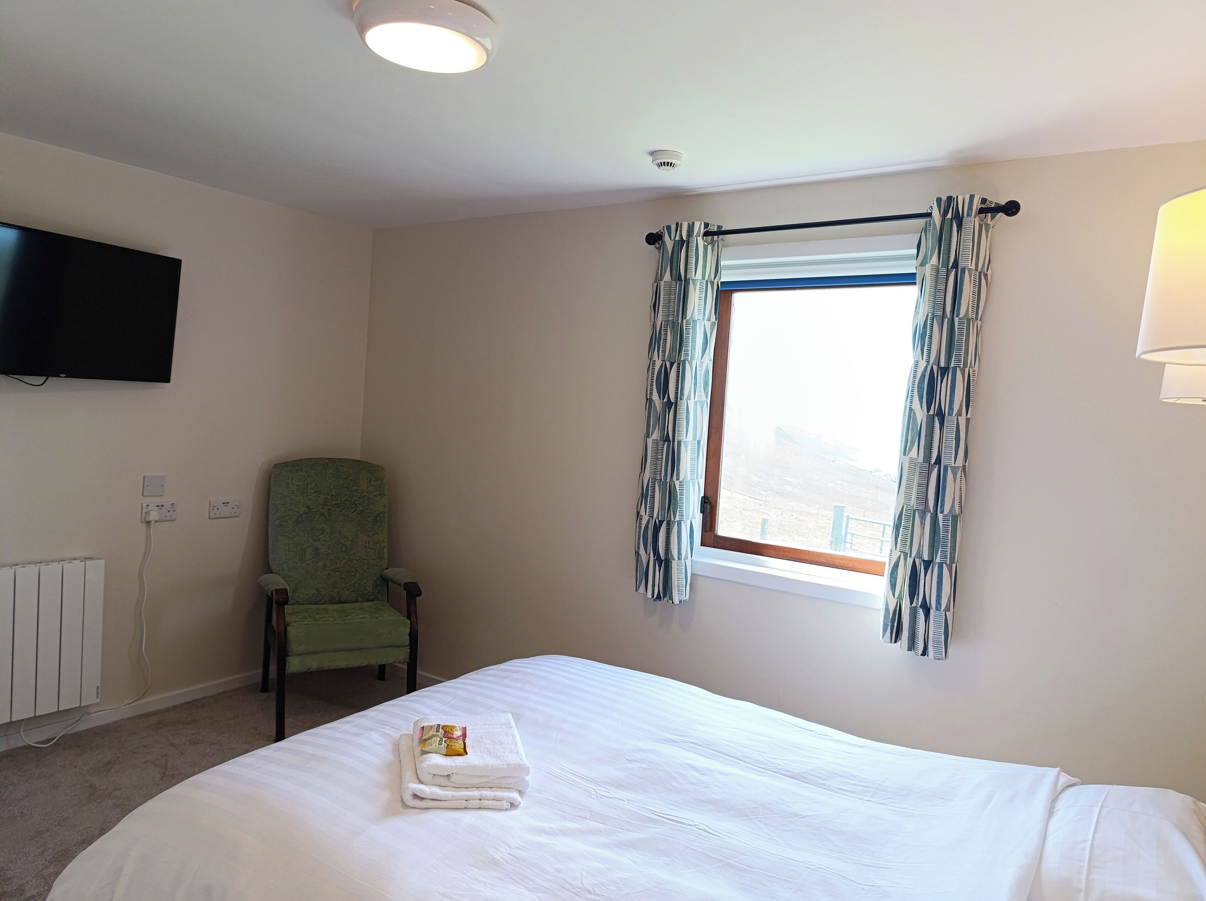 A hotel room with a bed, a chair, a window with curtains, a flat-screen TV mounted on the wall, and a ceiling light.