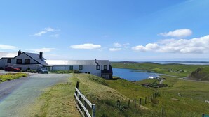 A coastal house with a driveway leading to a grassy area, overlooking a body of water and distant hills.