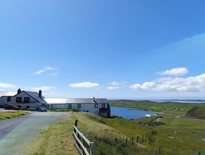 A coastal house with a driveway leading to a grassy area, overlooking a body of water and distant hills.