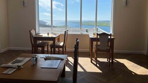 A room with a large window overlooking a coastal view, wooden furniture, and a tiled floor. - Westings Inn (Whiteness)