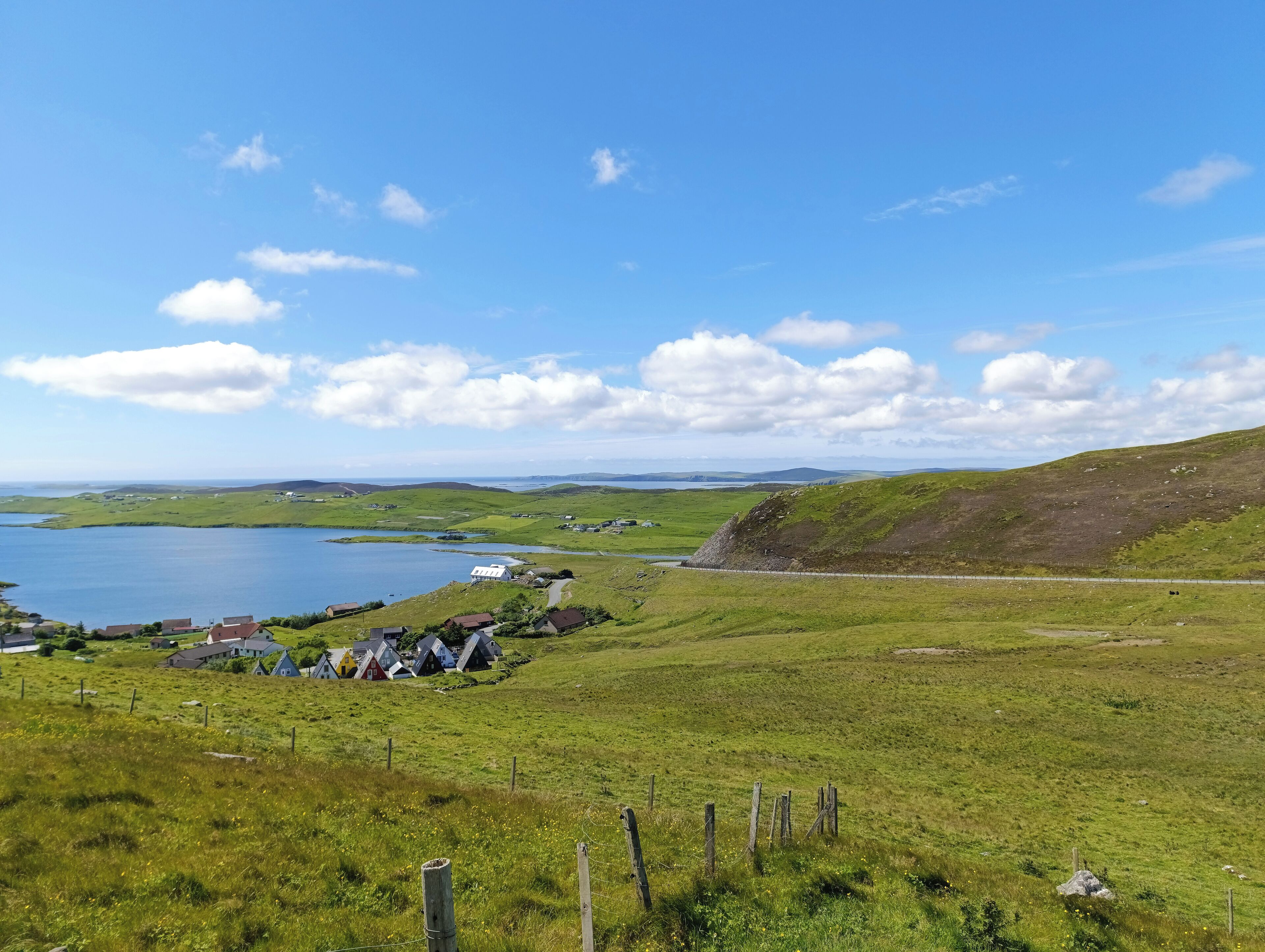 A coastal landscape with houses near a body of water, surrounded by green fields and hills.