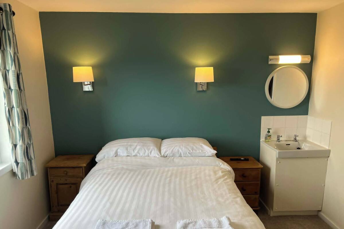 Une chambre à coucher comprenant un lit, deux lampes de chevet, un miroir rond, une table de chevet et une salle de bain avec un lavabo.