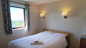 A neatly made bed with a white duvet and two pillows, a window with patterned curtains, and two wall-mounted lamps. - Westings Inn (Whiteness)