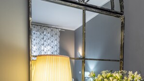 Un miroir avec un cadre noir, une lampe de table et un vase de fleurs posés sur une console blanche.