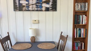 A dining table with a blue tablecloth, two woven placemats, wooden chairs, a wall-mounted sign, a wall-mounted lamp, and a bookshelf with books. - Newly renovated kitchenette. The Cozy cottage at the Marion County Lake. (Marion)