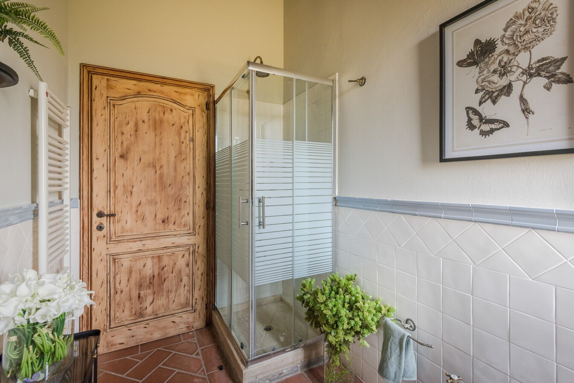 A wooden door, a glass shower enclosure, a tiled wall with a floral print, and a potted plant.