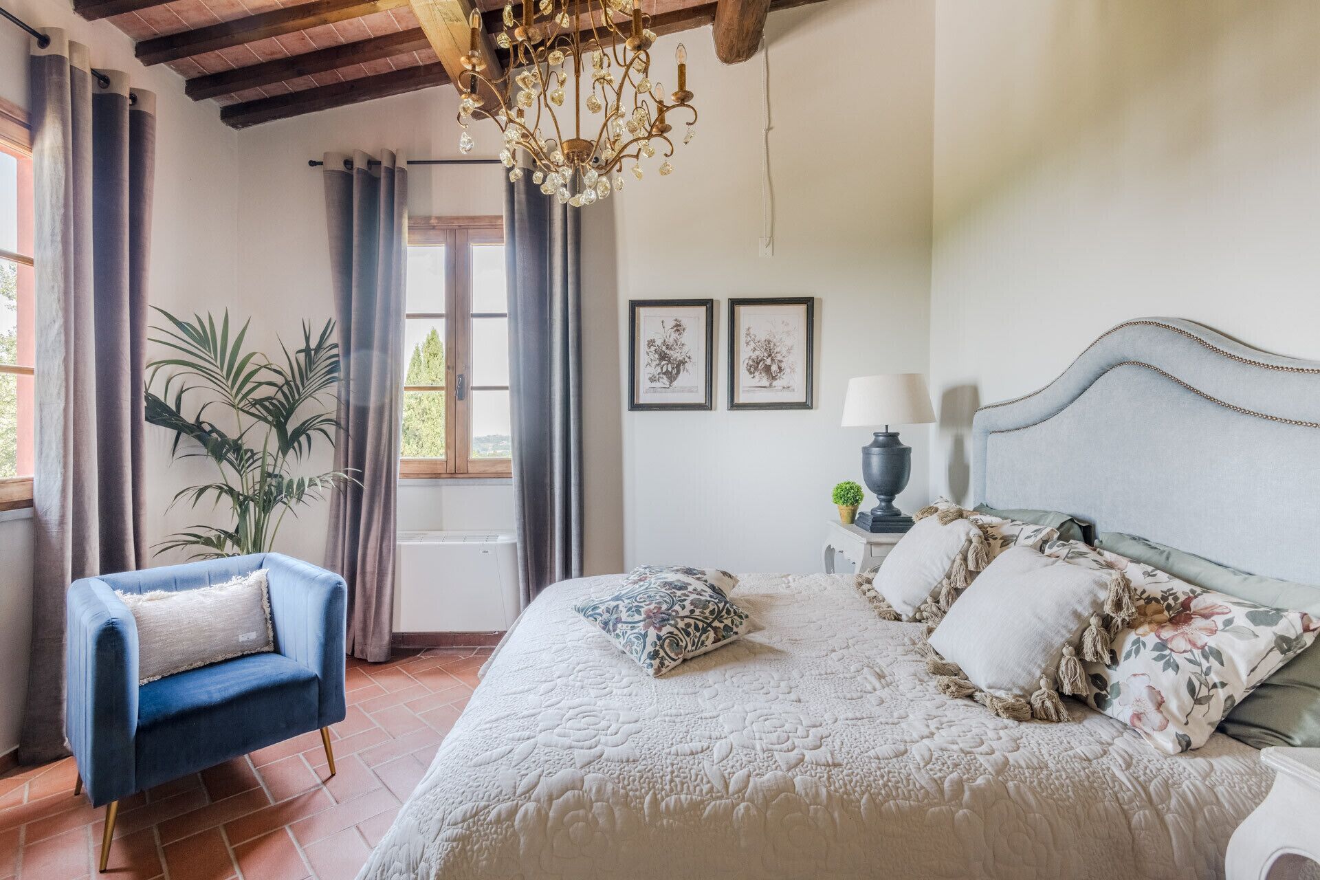 A bedroom with a large bed, a blue armchair, a chandelier, and two framed pictures on the wall.