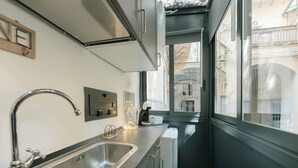 Eine schmale Küche mit einer Spüle aus Edelstahl, einer Kaffeemaschine und einem Fenster mit Blick auf einen Innenhof.