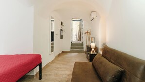 Ein schmaler Flur mit einem Bett links, einem braunen Sofa rechts und einer Treppe, die auf einen Balkon führt.
