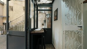 Ein enger Korridor mit einem Holztisch und Stühlen, einer Tür mit schmiedeeisernen Verzierungen und einer sichtbaren Treppe hinter der Glastür.