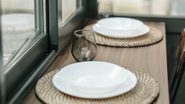 Ein Esstisch mit zwei weißen Tellern und einem Glas, gedeckt mit gewebten Platzsets, neben einem Fenster.