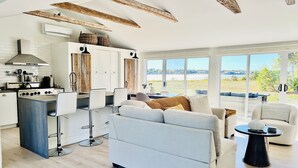 A spacious living area with a modern kitchen, large windows offering a view of the outdoors, and a cozy seating arrangement. - ✨ The Dream Cottage– Sea front w/ Hot Tub & Sunset Views - near Parlee Beach (Shediac)