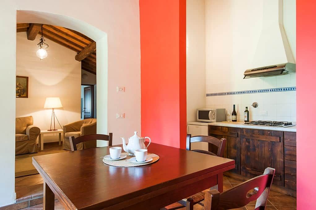 A dining area with a wooden table set for two, a kitchen with a microwave and stove, and a living area with a sofa and lamp.