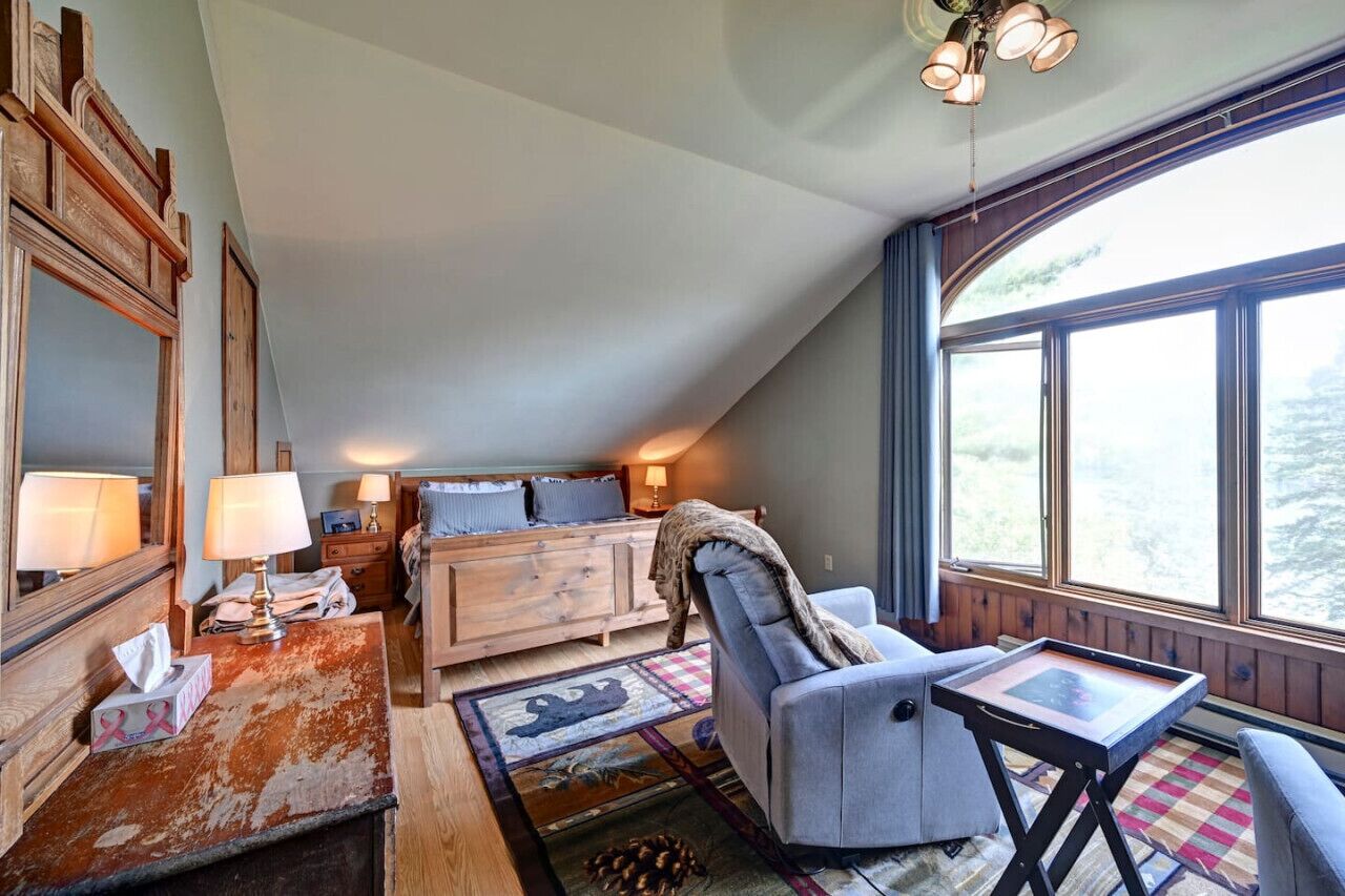 A cozy room with a wooden dresser, a sofa, a chair, and a small table. There is a large window providing a view of the outdoors.