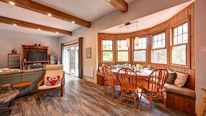 A cozy cabin interior with a dining area, a living room with a sofa and TV, and large windows providing natural light.