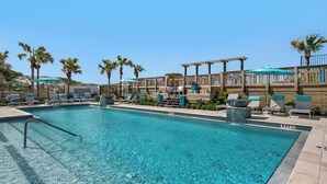 Une piscine d’un bleu éclatant, bordée de chaises longues et d’ombrelles, entourée de palmiers et d’une terrasse en bois.
