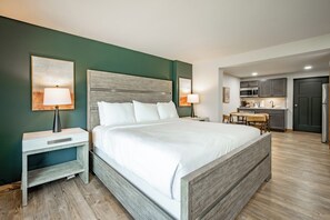 A modern hotel room with a large bed, a nightstand, a desk, and a kitchenette.