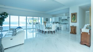 Un espacio habitable amplio con un gran ventanal que da a un balcón, una zona de comedor con mesa de vidrio y una cocina al fondo.