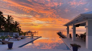 Zona de piscina frente al agua con sillones de descanso y un área de estar cubierta, con vista a un mar tranquilo al atardecer.