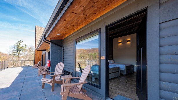 A modern log cabin with a patio area featuring Adirondack chairs, a fire hydrant, and a large sliding glass door leading to a bedroom with a bed and a nightstand. - Cove Creek Lodge: 203 Poolside Double Queen Room (Oakland)