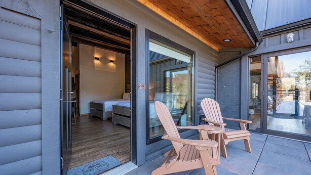A modern log cabin with a wooden deck, two Adirondack chairs, and a cozy interior with a bed and a small table. - Cove Creek Lodge: 205 Poolside Double Queen Room (Oakland)