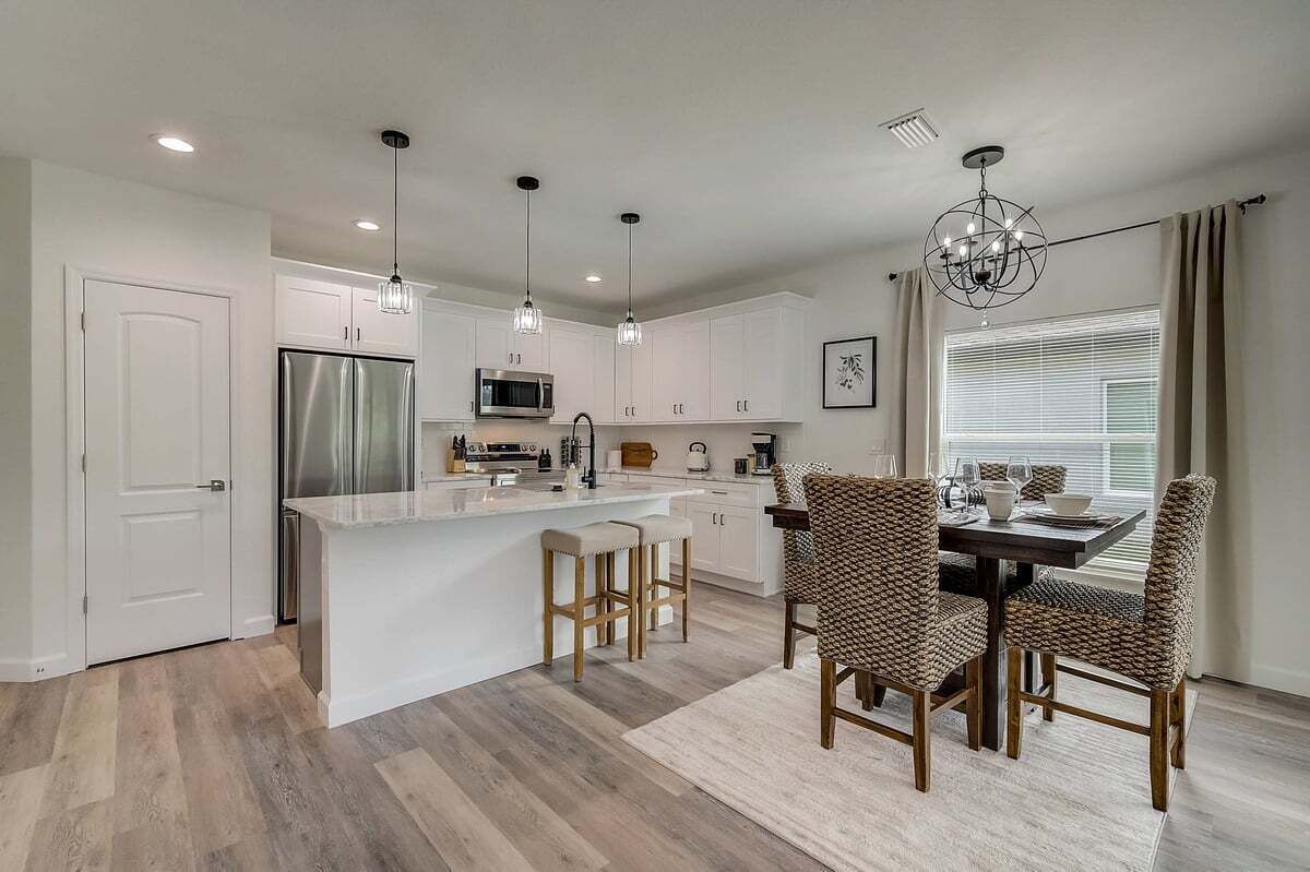 Una cocina moderna con armarios blancos, un refrigerador de acero inoxidable, una isla central con taburetes, iluminación colgante y un comedor con sillas de mimbre.
