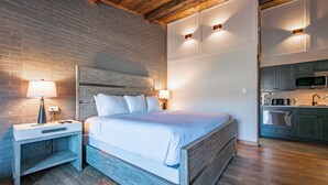 A modern bedroom with a wooden headboard, a nightstand with a lamp, and a built-in kitchen area with a microwave and sink.