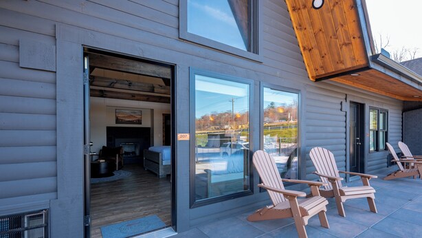 Une cabane en rondins moderne, dotée d’une entrée avec une porte en verre, d’une chambre visible avec un lit et un bureau, et de deux fauteuils Adirondack sur la terrasse.