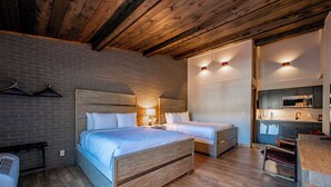 A modern hotel room with a wooden ceiling, a brick accent wall, a large bed with a headboard, a small kitchenette, and a seating area with a chair.