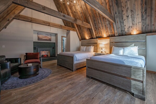 A spacious loft bedroom with a high ceiling, exposed wooden beams, a fireplace, two beds, a sofa, and a coffee table.
