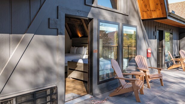 A modern building with a large glass door entrance, wooden deck chairs, and a visible bed inside. - Cove Creek Lodge: 217 Lakeside Double Queen Fireplace Room (Oakland)