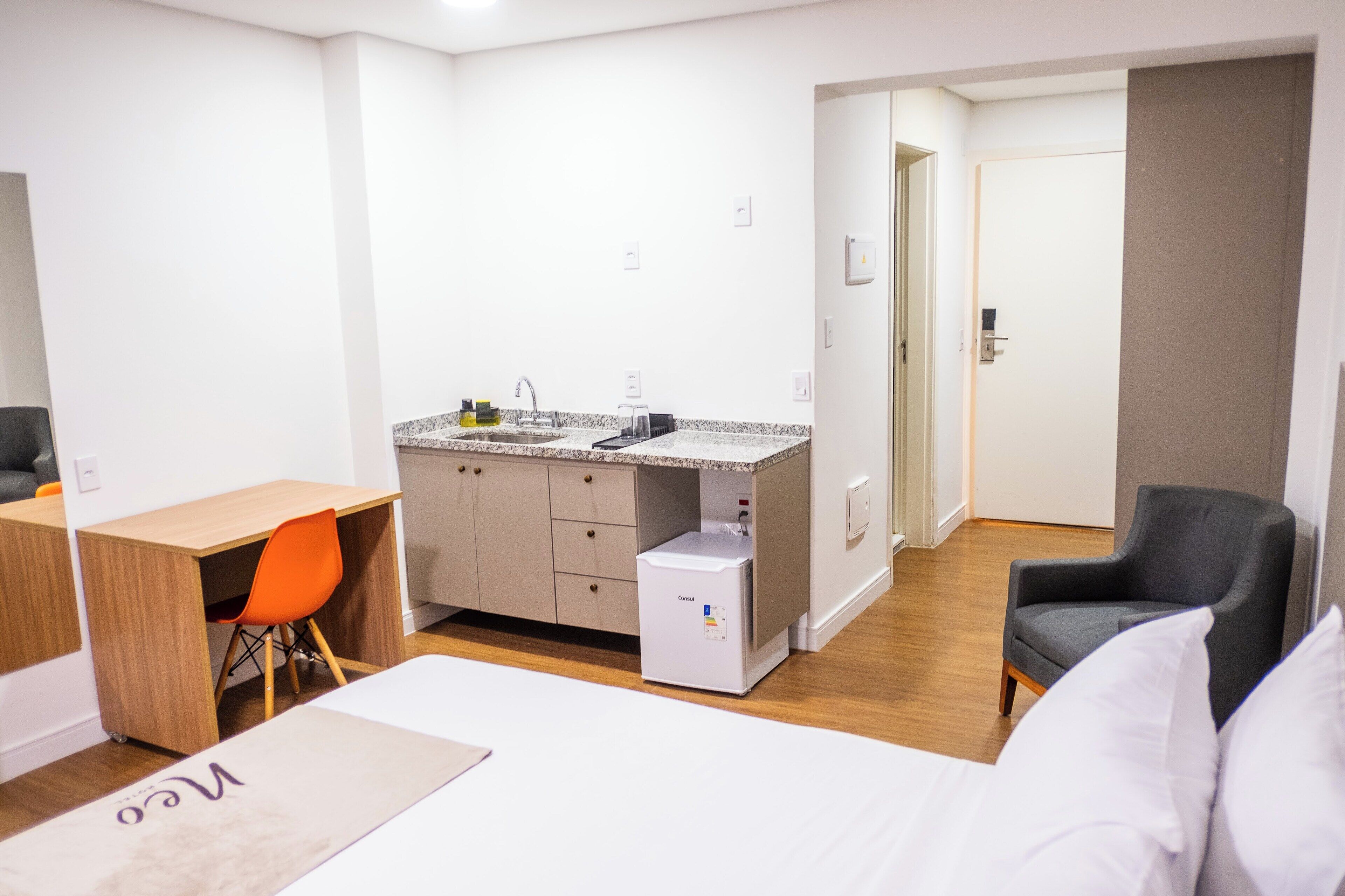 A compact hotel room with a bed, a desk, a chair, a kitchenette, and a separate bathroom.