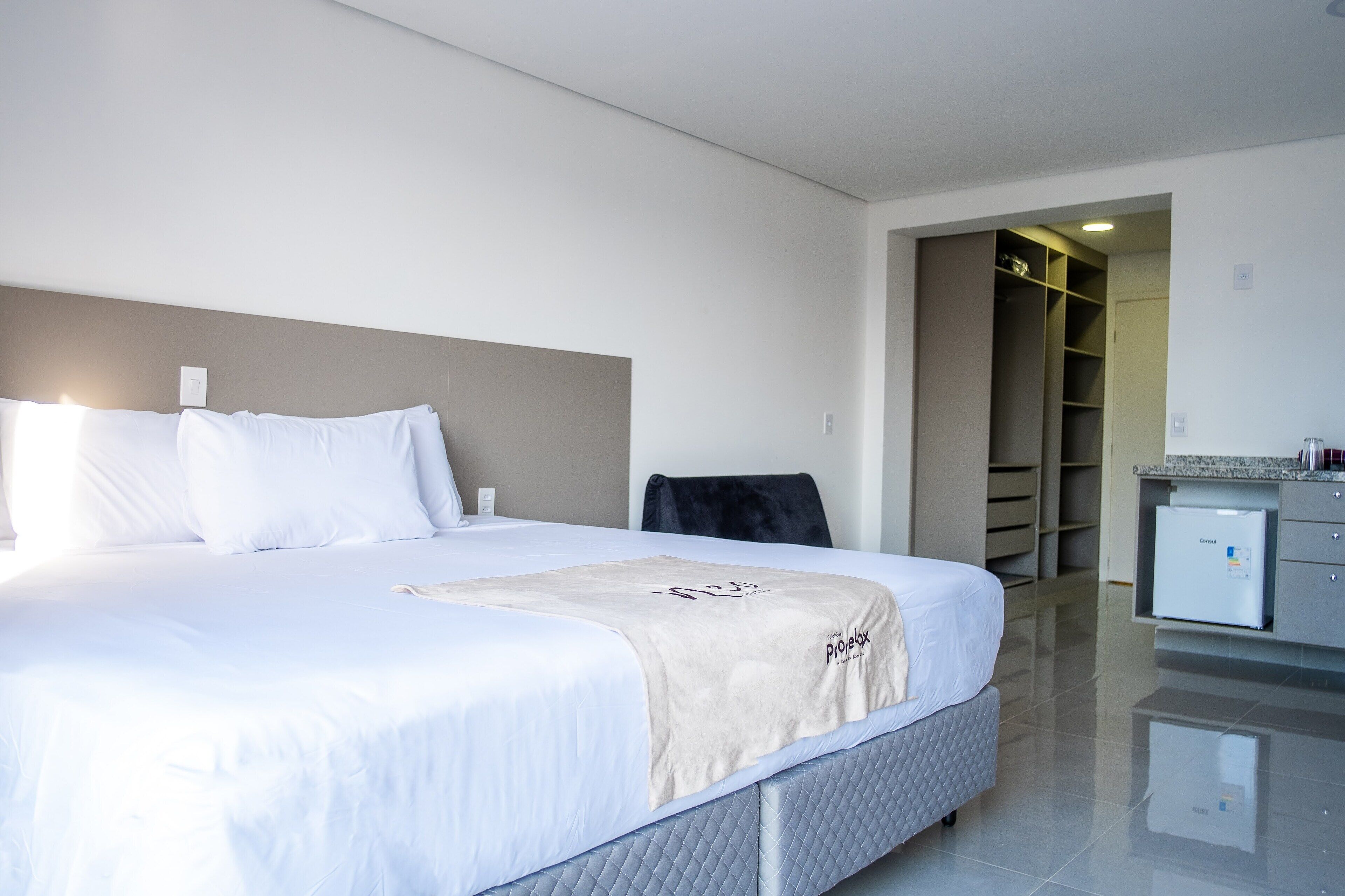Una habitación de hotel con una cama grande, una mesita de noche con refrigerador y un vestidor.