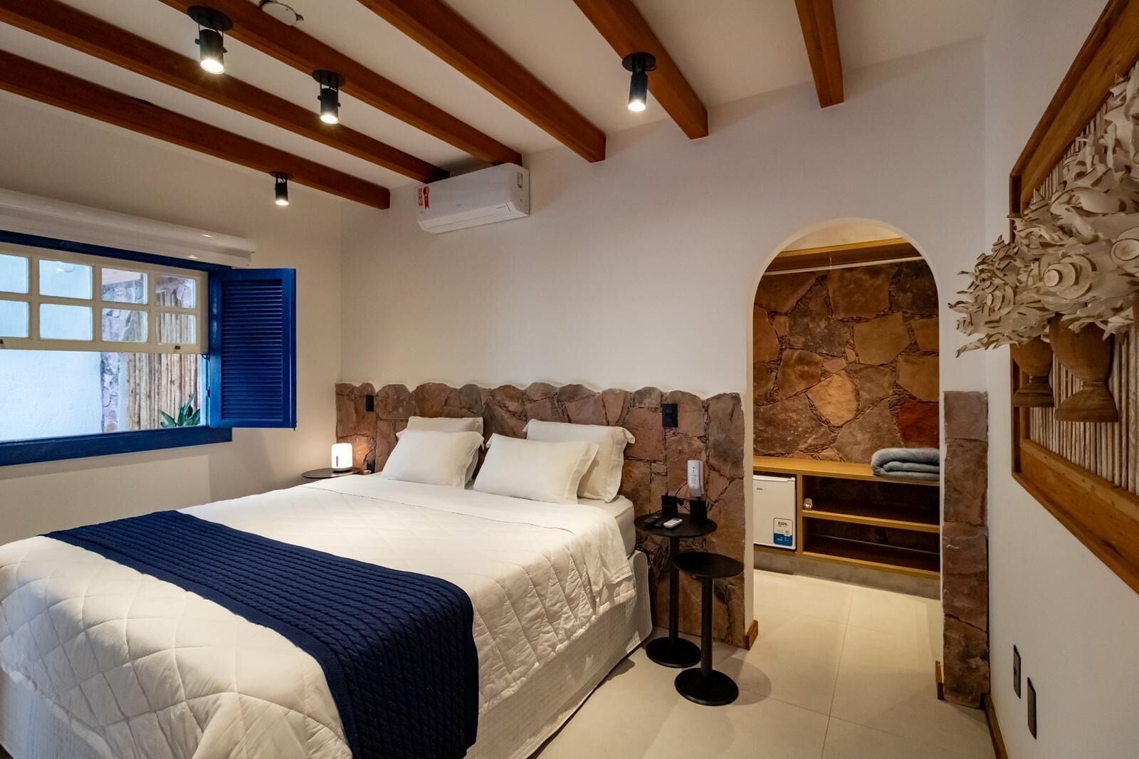 Um quarto com uma cama grande, uma parede de pedra, uma mesinha com abajur e uma janela com persianas azuis.