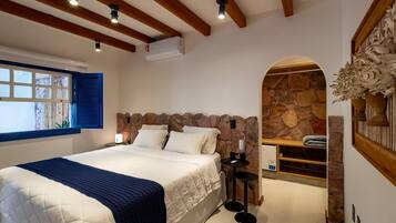 Um quarto com uma cama grande, uma parede de pedra, uma mesinha com abajur e uma janela com persianas azuis.