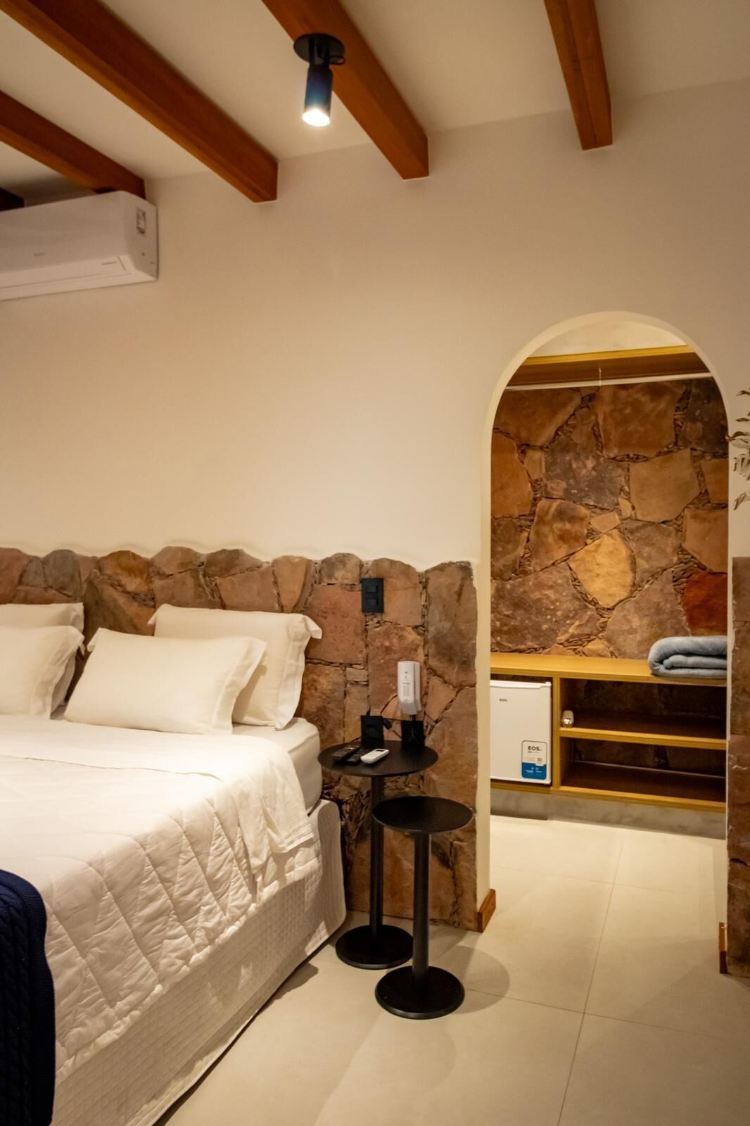 Un dormitorio con pared de piedra, una cama con ropa de cama blanca, una mesita con un control remoto y un estante de madera con toallas.