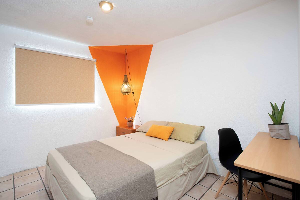 Ein Schlafzimmer mit Bett, Schreibtisch, Stuhl, einer Topfpflanze und einem Fenster mit Jalousie.