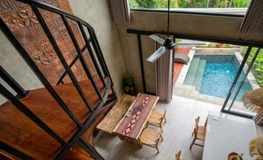 A modern interior with a wooden staircase, a dining area with a patterned tablecloth, and a view of an outdoor swimming pool. - Juminten Villa  (Ubud)