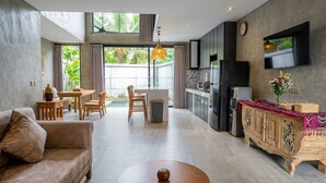 A modern living room with a sofa, dining table, and chairs, a kitchen with a refrigerator and microwave, and a large window with a view of greenery. - Juminten Villa  (Ubud)