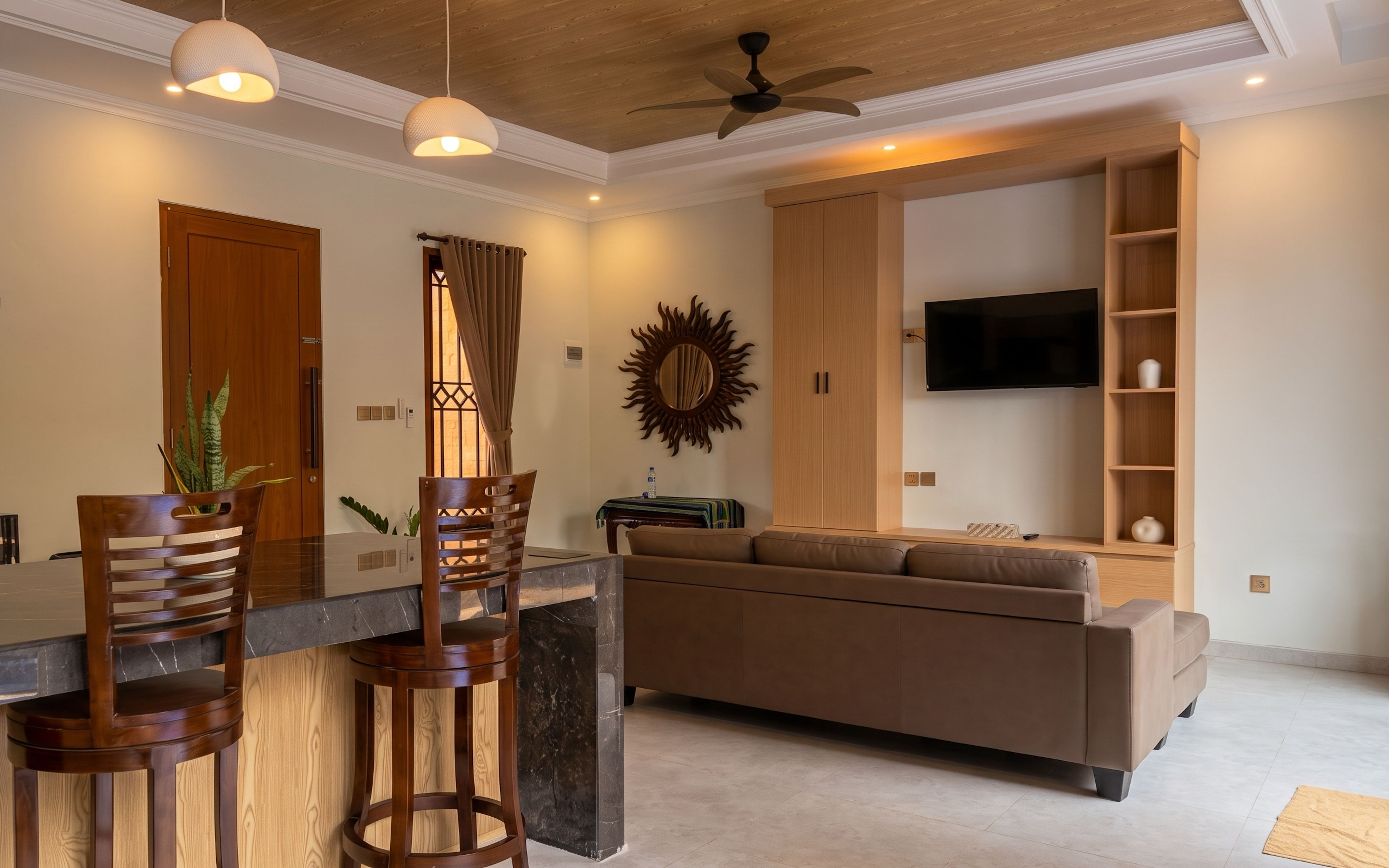 A modern living room with a sofa, a dining area with chairs and a table, a TV mounted on the wall, and a ceiling fan.