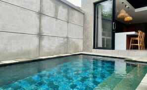 A swimming pool with clear blue water, a modern concrete wall, and a glass door leading to an interior space with a dining area. - Juminten Villa  (Ubud)