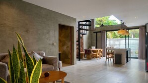 A modern living room with a sofa, coffee table, and a large plant. There is a dining area with a table and chairs, and a kitchen area with a stove and sink. - Juminten Villa  (Ubud)